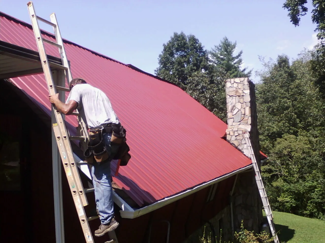 Roofer on a ladder beside a red metal roof and stone chimney — Soffit & Fascia Repair by Roofing Brothers in Kennard